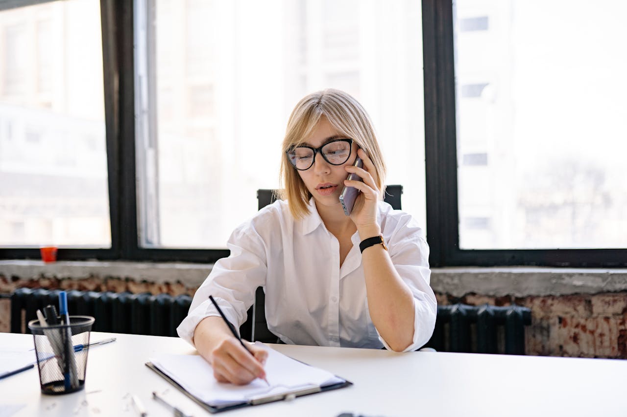 A woman in an office setting multitasking with a phone call and taking notes.